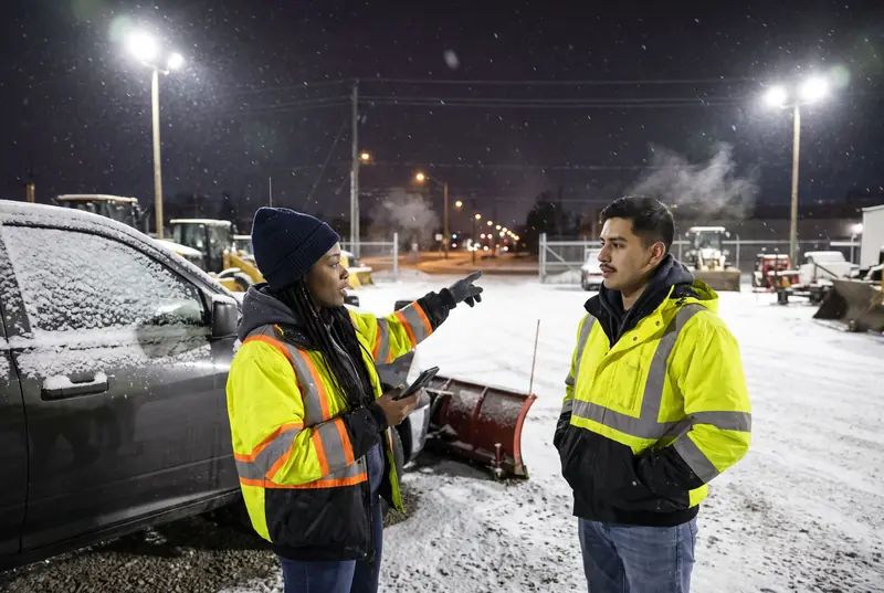 Two crew members in hi-vis jackets talking beside a plow truck in a yard at night